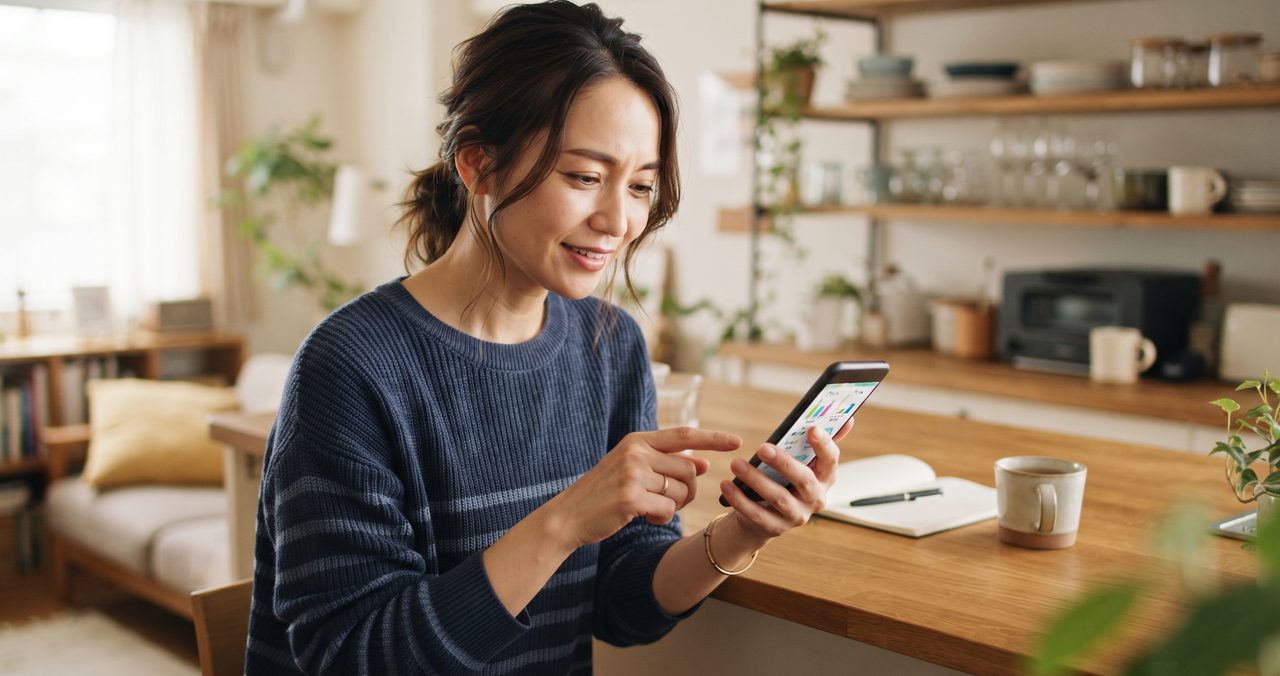 自宅のキッチンでスマートフォンの通話プラン比較画面(無地)を指で操作しながら確認している女性