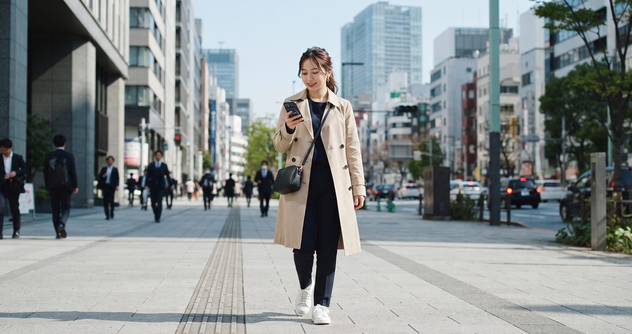 オフィス街の歩道を歩きながらスマートフォンの画面を見ている女性の全身ショット、電波の繋がりやすさをイメージした情景