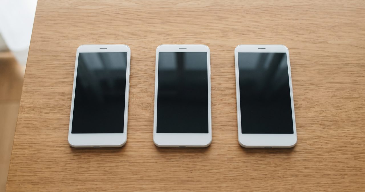 木製テーブルに並べられた無地のスマートフォン3台、ドコモ・au・ソフトバンク3回線から選べるイメージ