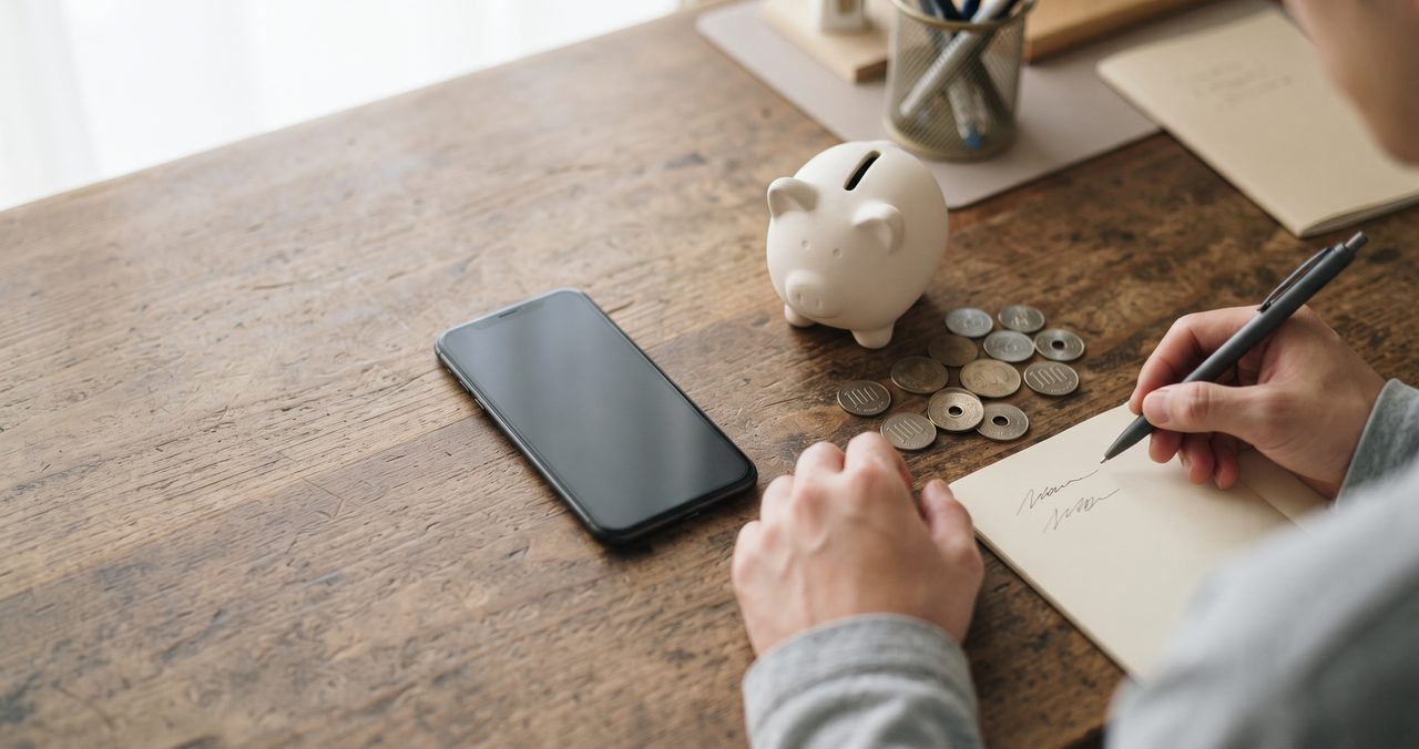 スマートフォンと貯金箱・硬貨を並べ、買い替え費用を考えている人物の手元