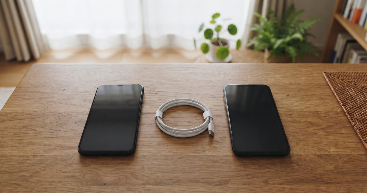 旧端末と新端末を並べて置き、その間に丸めた充電ケーブルを置いた木製テーブルの上の俯瞰の物撮り